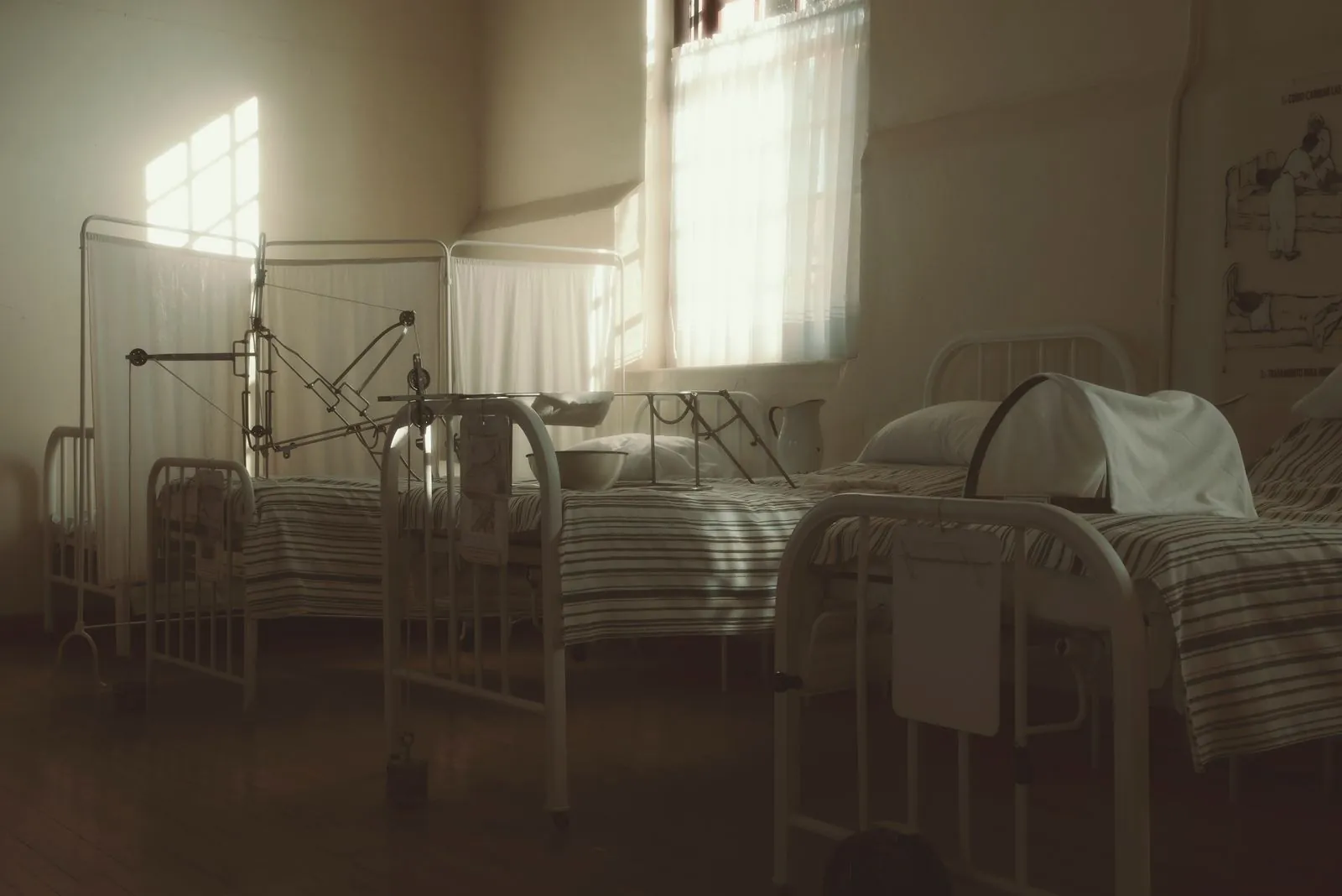 Salle d’hôpital ancienne faiblement éclairée, avec des lits alignés et du matériel d’époque.